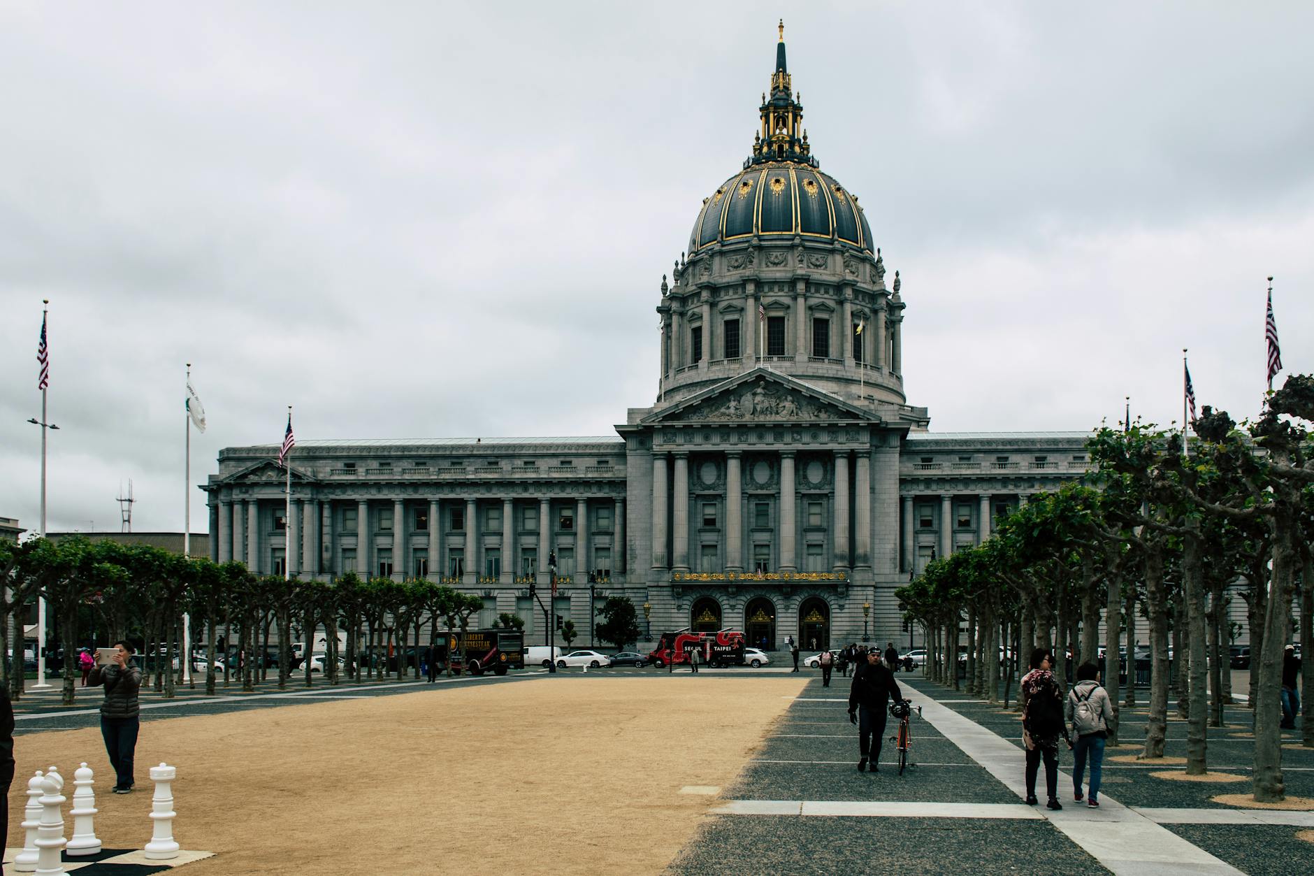 A view of San Francisco City Hall with a public square, trees, and people under a cloudy sky.